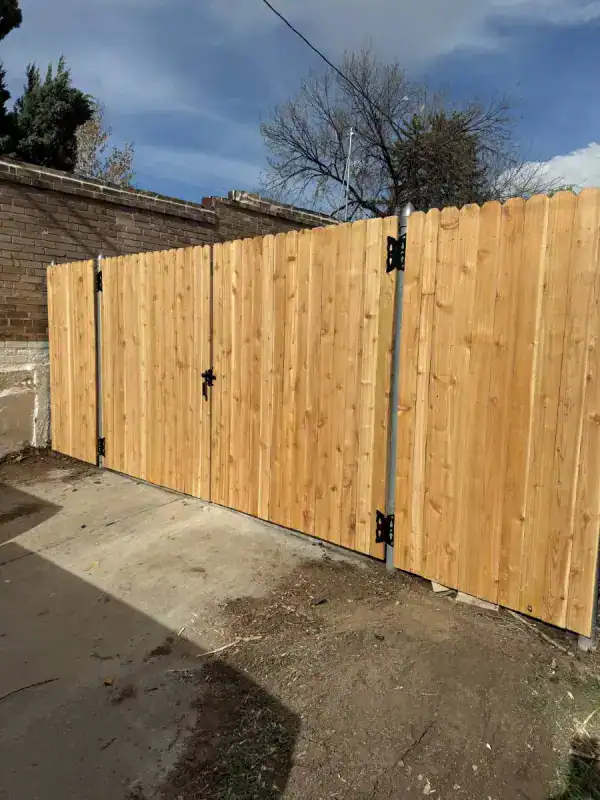 New light-wood privacy fence with metal posts along a concrete ground, brick wall behind and leafless trees against a blue sky.